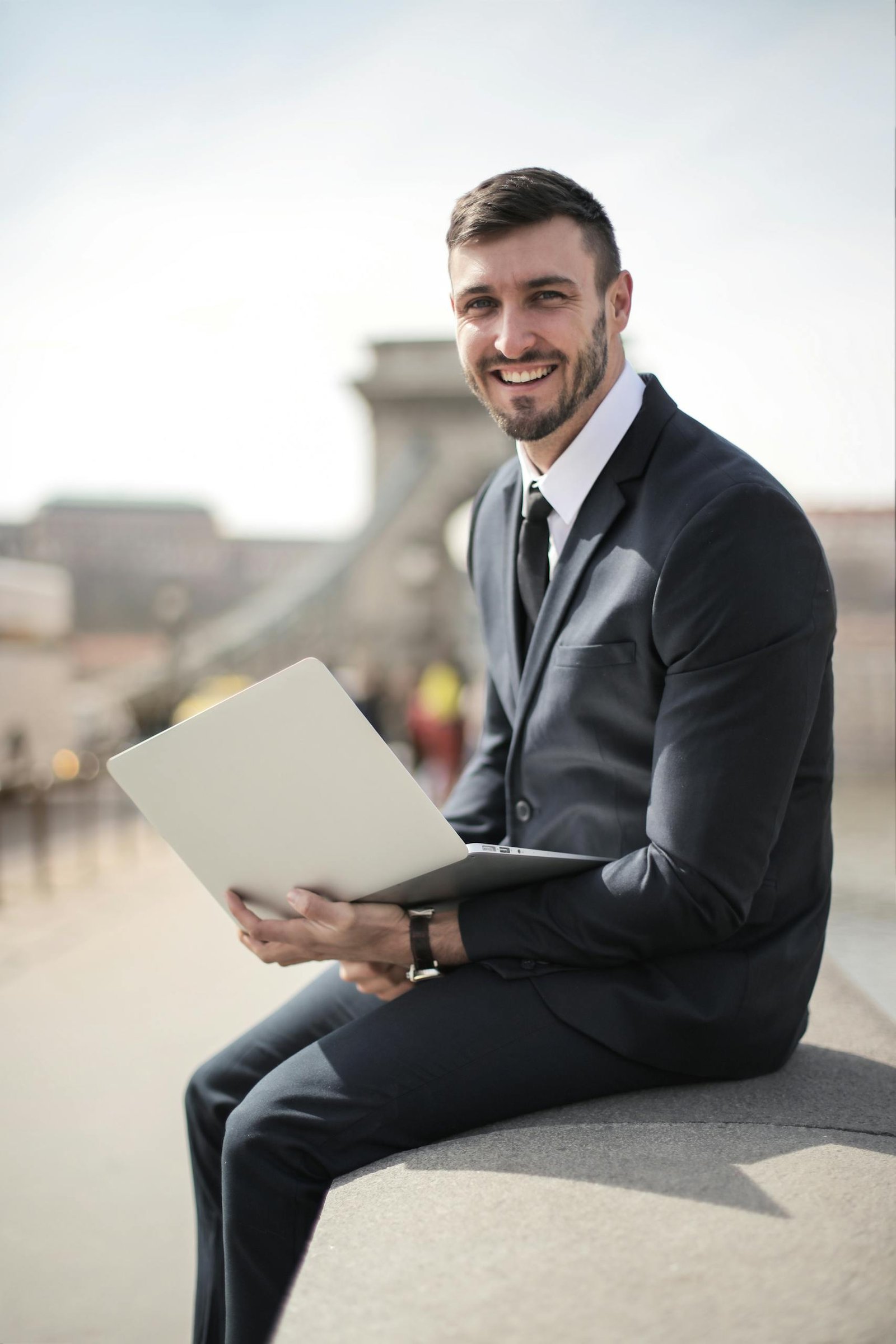 Happy businessman in suit holding laptop while sitting on a city bridge, showcasing modern corporate lifestyle.