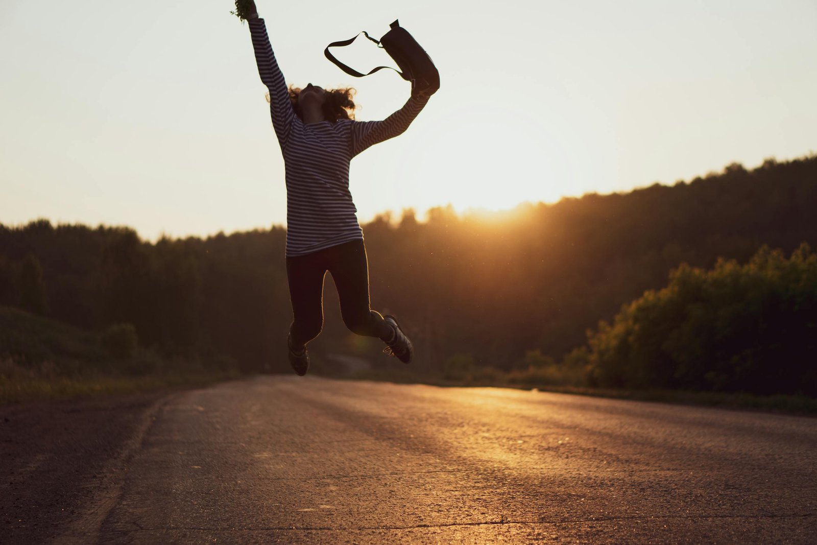 Silhouette of a woman jumping joyfully on an open road at sunset, embodying freedom and adventure.